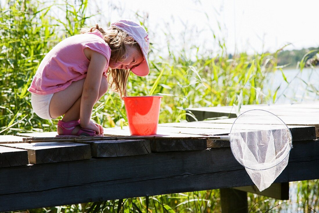 Little girl with fishing net looking into bucket on wooden jetty