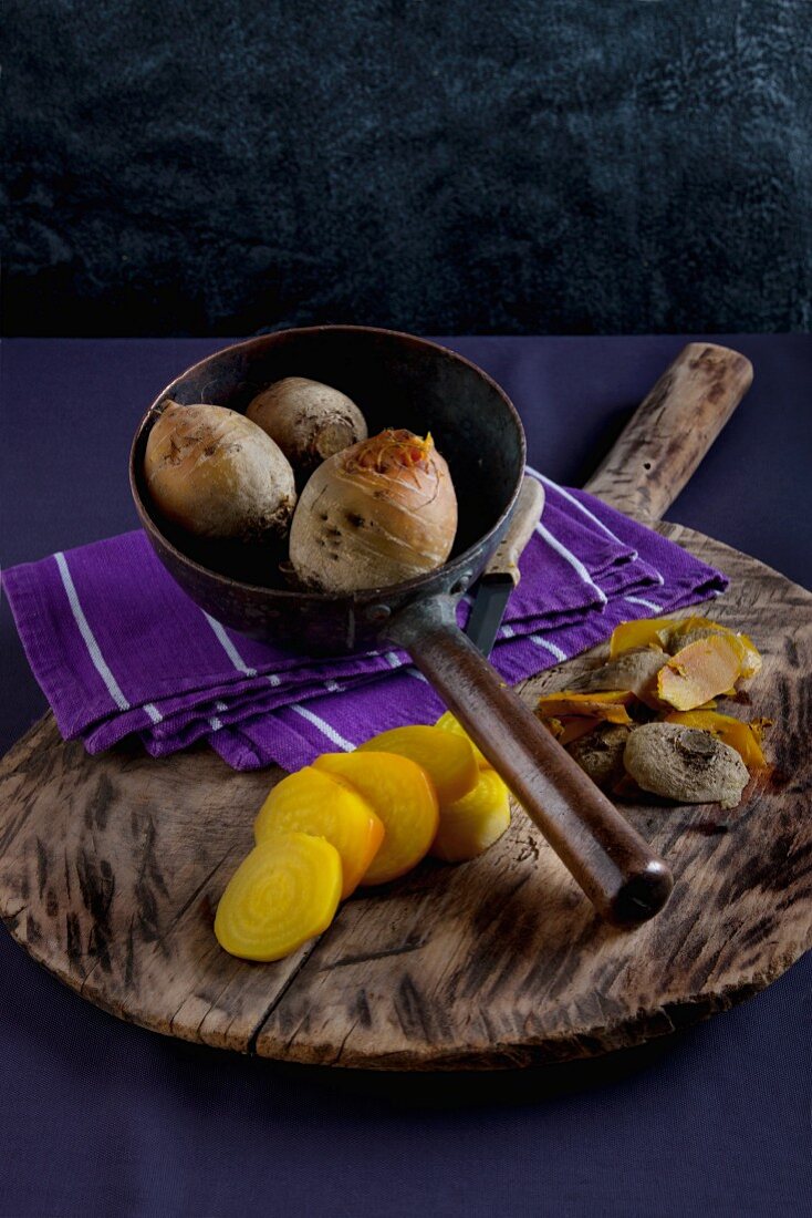 Gelbe Bete, gekocht, teilweise geschält, in Scheiben geschnitten, auf Holzbrett und in alter Metallkelle mit Tuch und Messer