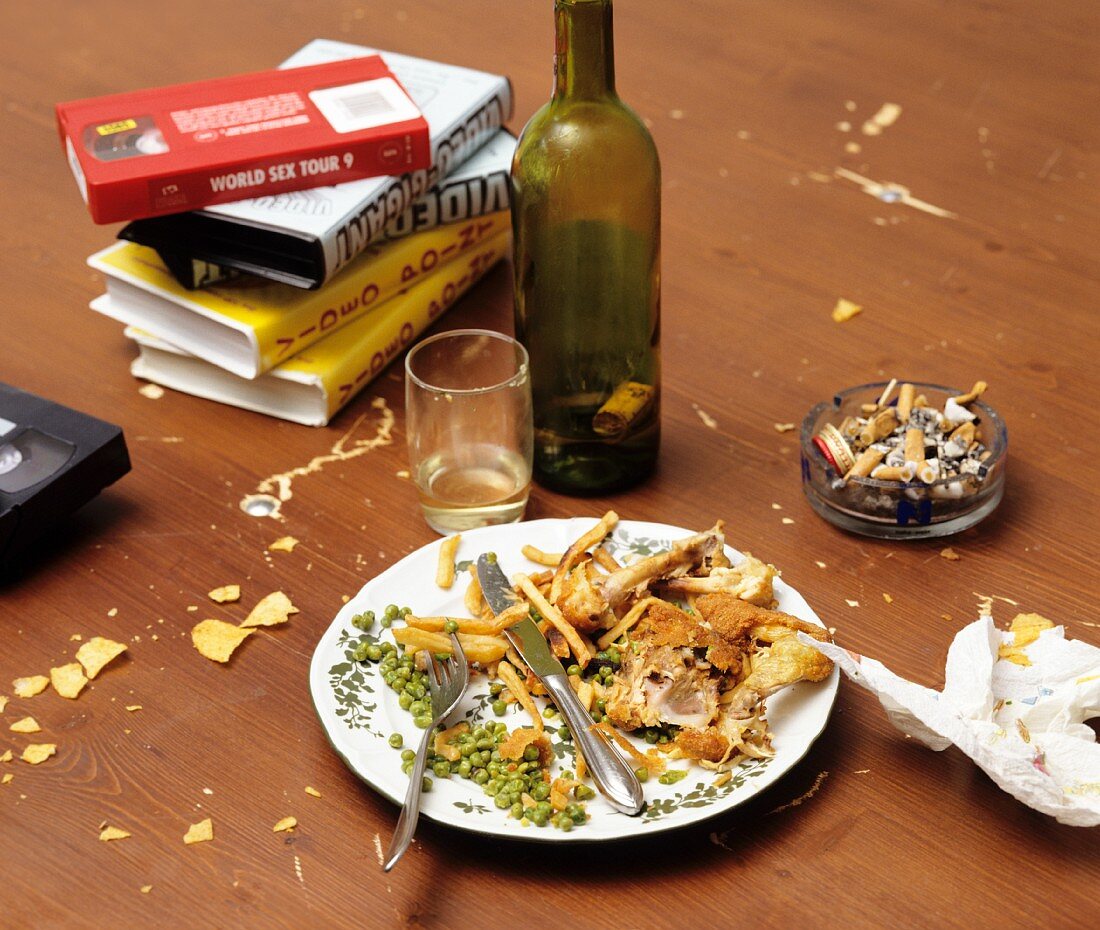 Remnants of a meal on a plate, an empty wine bottle, a full ashtray and video tapes