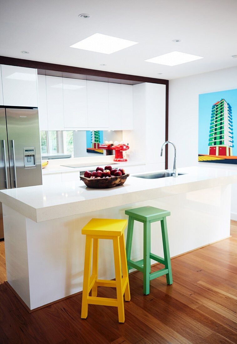 Coloured wooden bar stools in front of white kitchen island with sink in open-plan designer kitchen