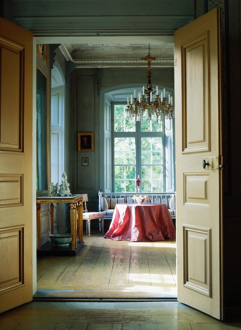 Open double doors showing view of table with pink tablecloth and bench in front of tall window in living room of stately home