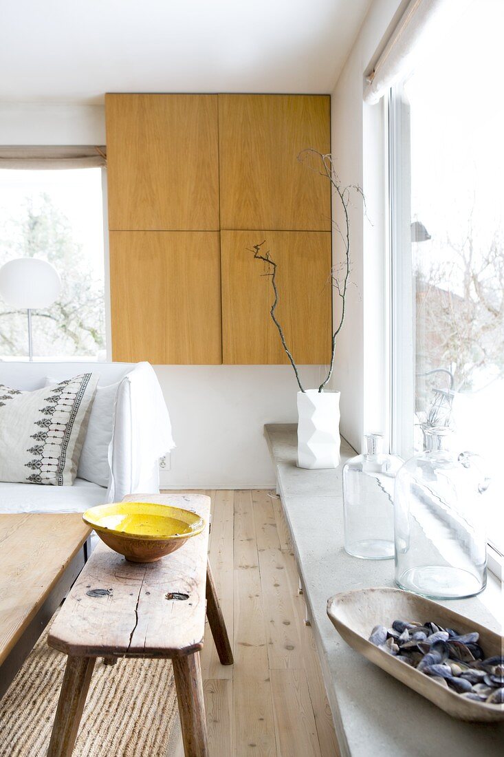 Fitted cupboard at end of window sill decorated with glass vessels next to ceramic bowl on old wooden bench