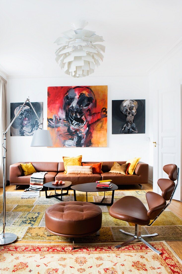 Classic office chair with brown leather cover and footstool in front of lounge area with black coffee table and brown leather sofa below expressive paintings on wall