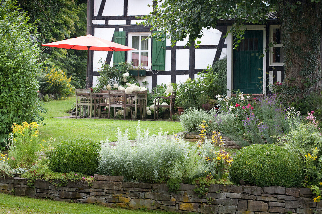 Seating area with parasol in front of renovated, old, half-timbered house in idyllic garden with low, dry stone wall
