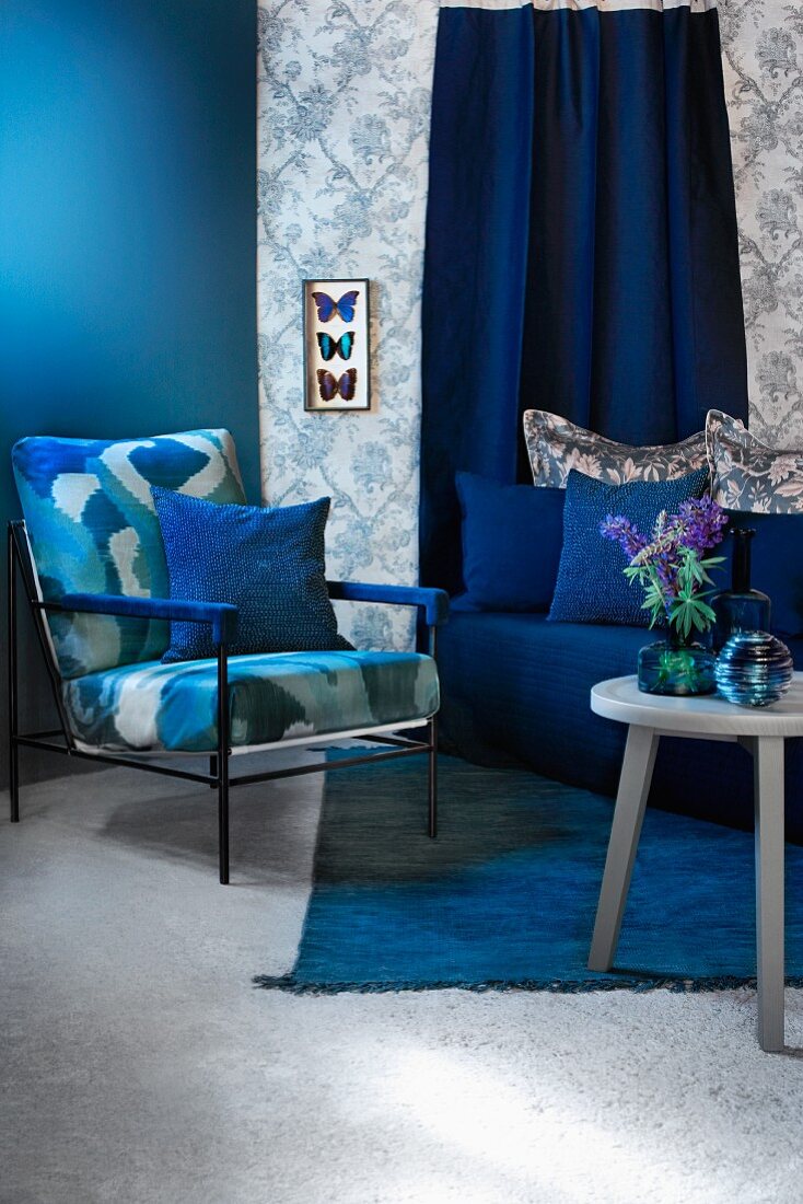 Sitzecke in Blautönen mit Polstersessel, Sofa und Beistelltisch; Schaukasten mit präparierten Schmetterlingen an Wand mit gemusterter Tapete