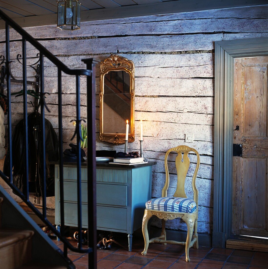 Schmiedeeisernes Treppengeländer; dahinter eine blaue Kommode mit goldgerahmtem Spiegel an rustikaler Holzwand
