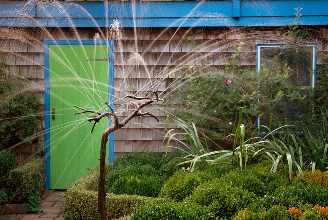 Original fountain with stylised tree branches in garden; wooden house with shingle facade in background