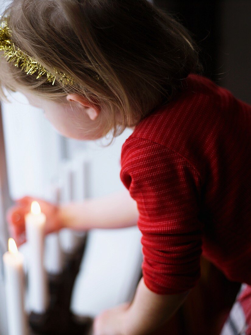 Little girl with tinsel wreath in hair looking at Christmas candles on windowsill