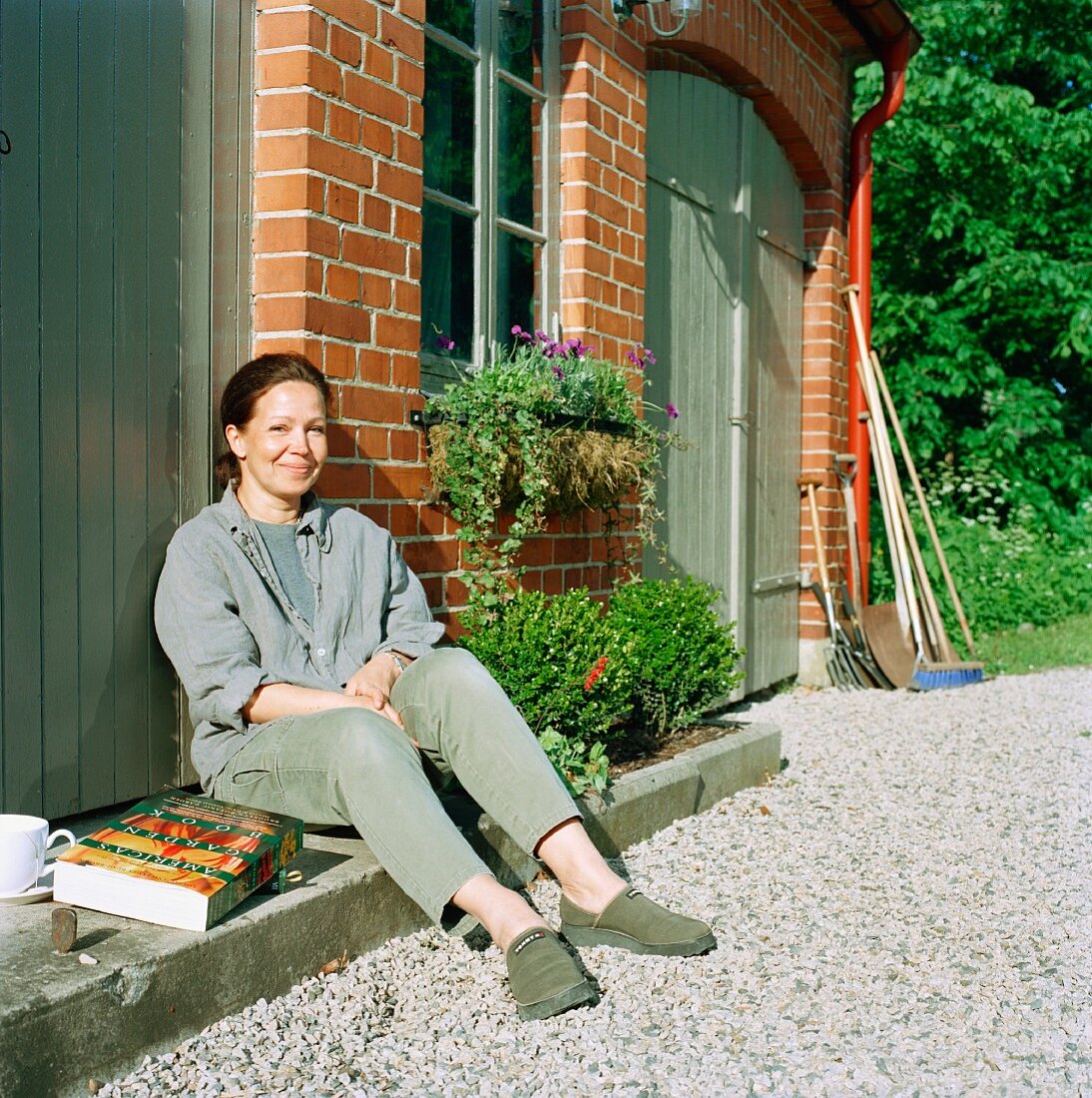 Woman with book & cup of coffee sitting on doorstep in sunny courtyard