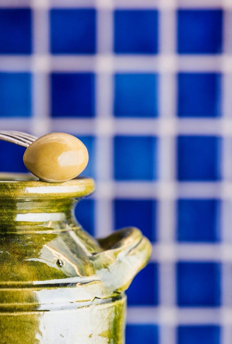 Fork holding green olive on top of a green ceramic jar. Blue tiles in the background.