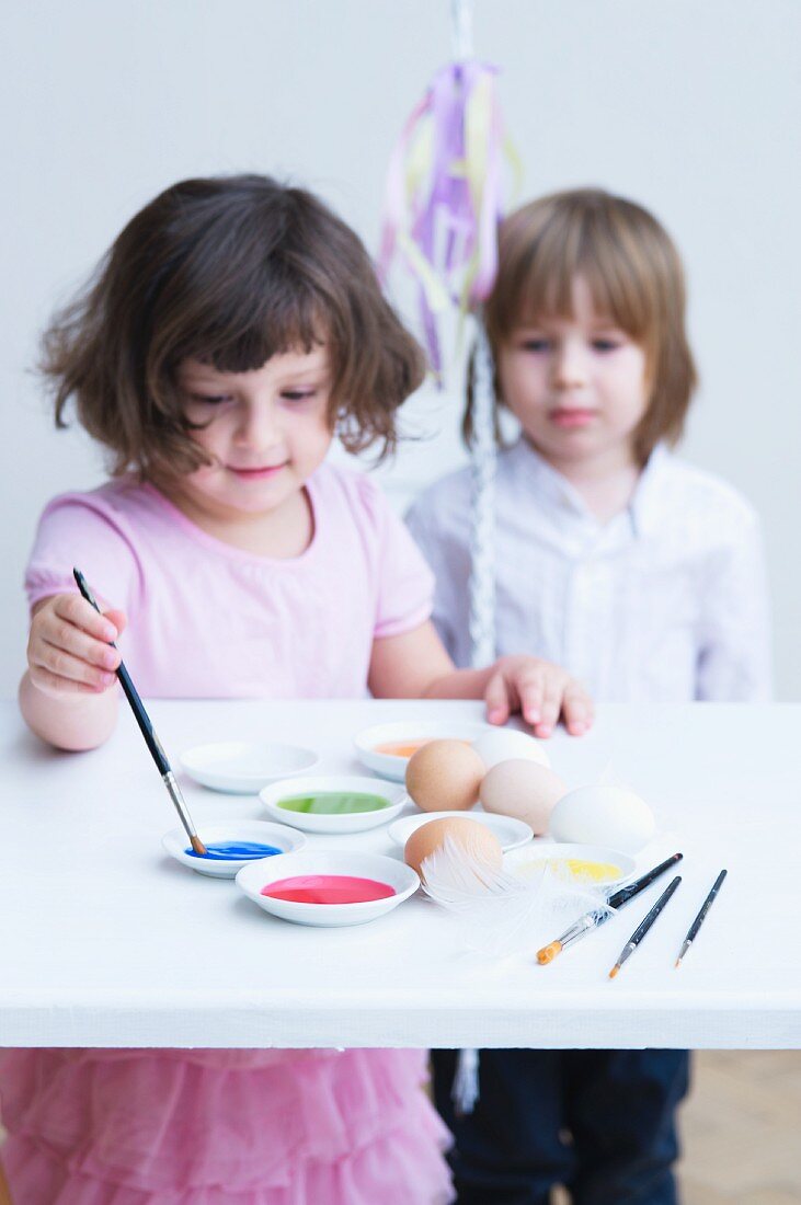 Little girl painting Easter eggs with second child watching