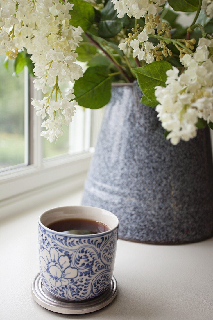 A mug of tea in front of a bunch of lilac flowers in a ceramic mug