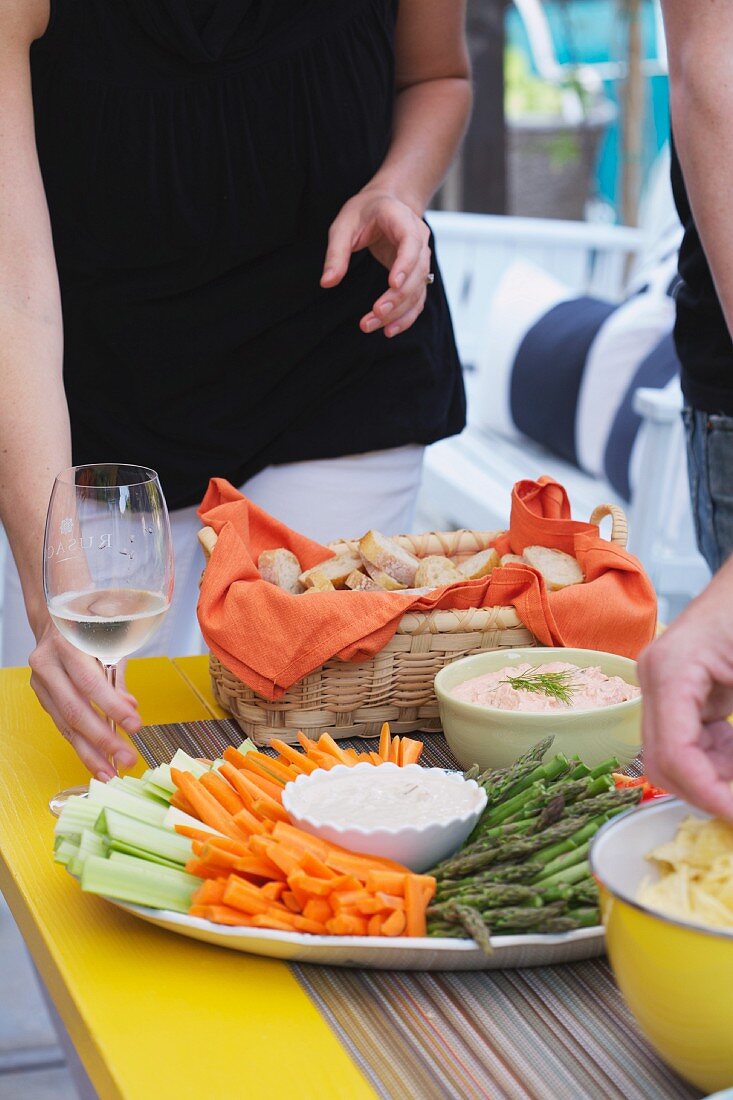 People by a buffet table set with a bread basket, a plate of crudités, and dips