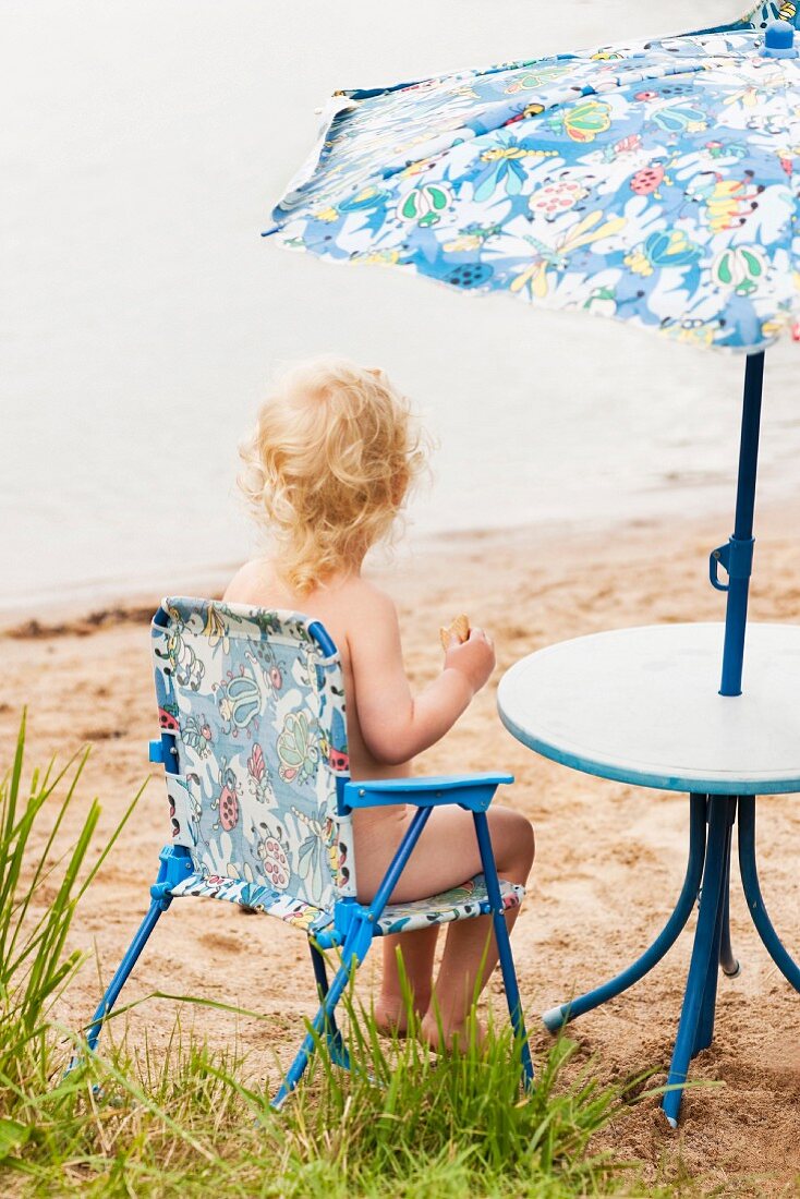 Kleinkind sitzt in Campingstuhl unter Sonnenschirm am Strand (Södermanland, Schweden)