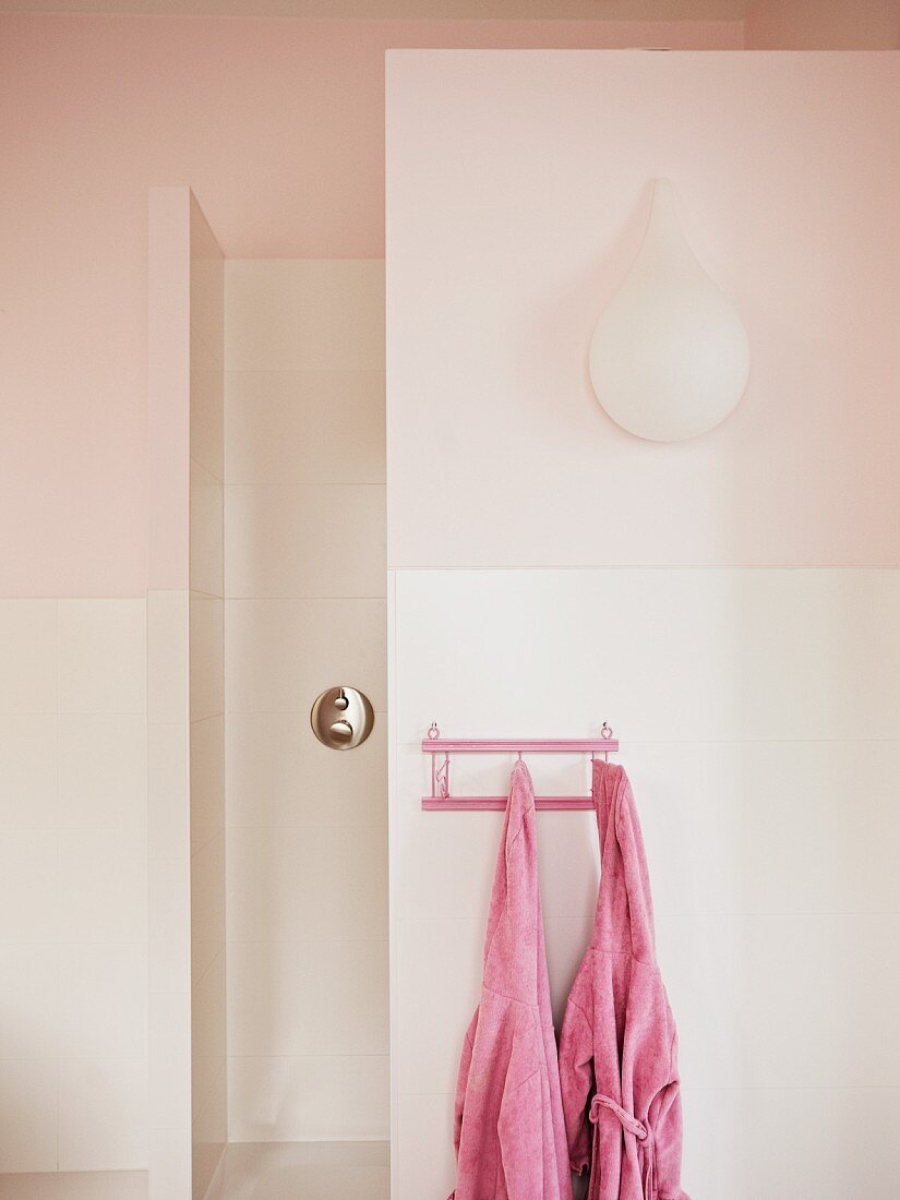 Teardrop-shaped sconce lamp on pink bathroom wall; two pink children's bathrobes hanging on small peg rack