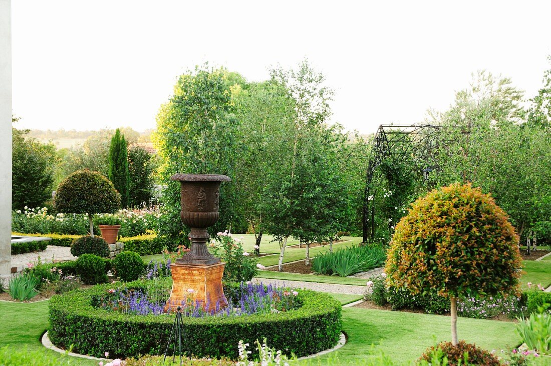 Landscaped garden with paths and clipped box hedges; central large urn in bed of summer flowers