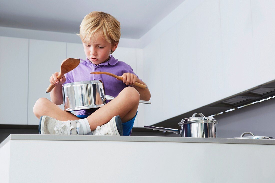 Boy sitting on kitchen counter playing drums with pans and wooden spoons