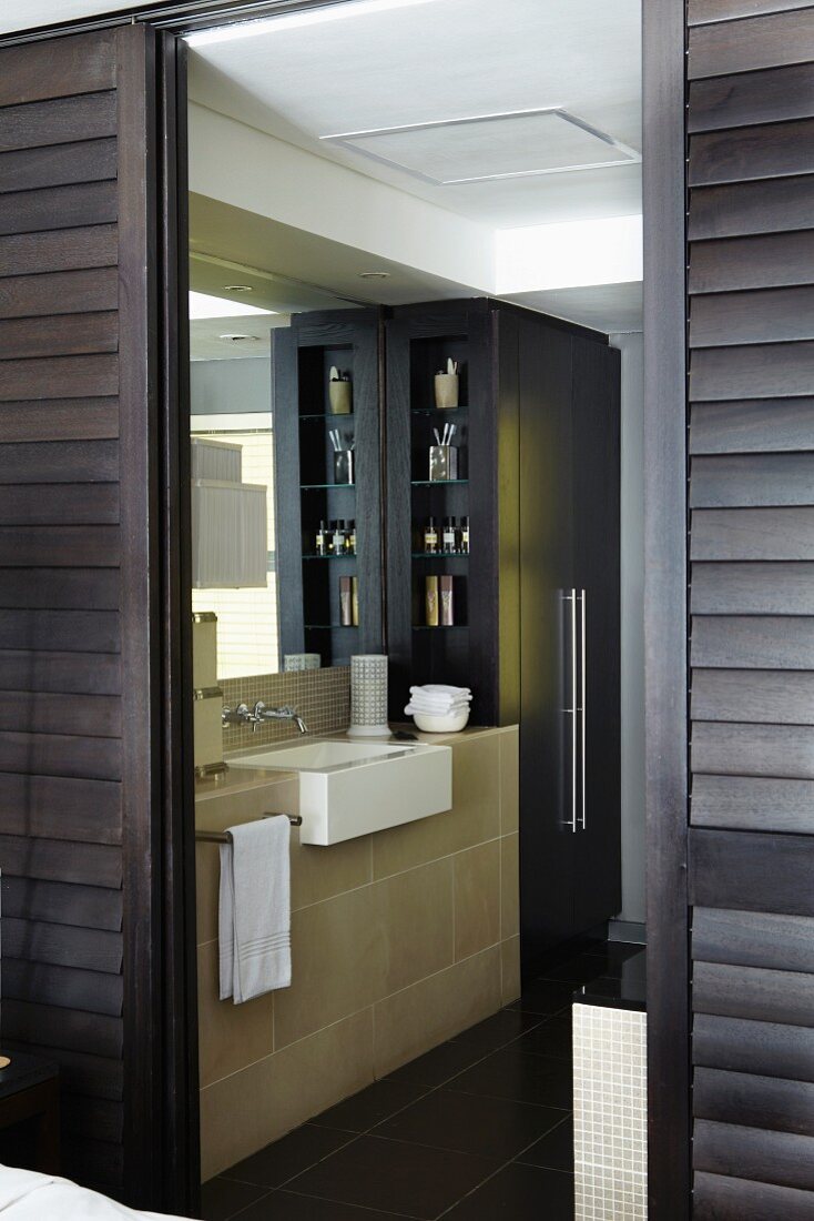 Masculine ensuite bathroom behind open, dark brown, sliding louver doors with elegant washstand and black fitted cupboard