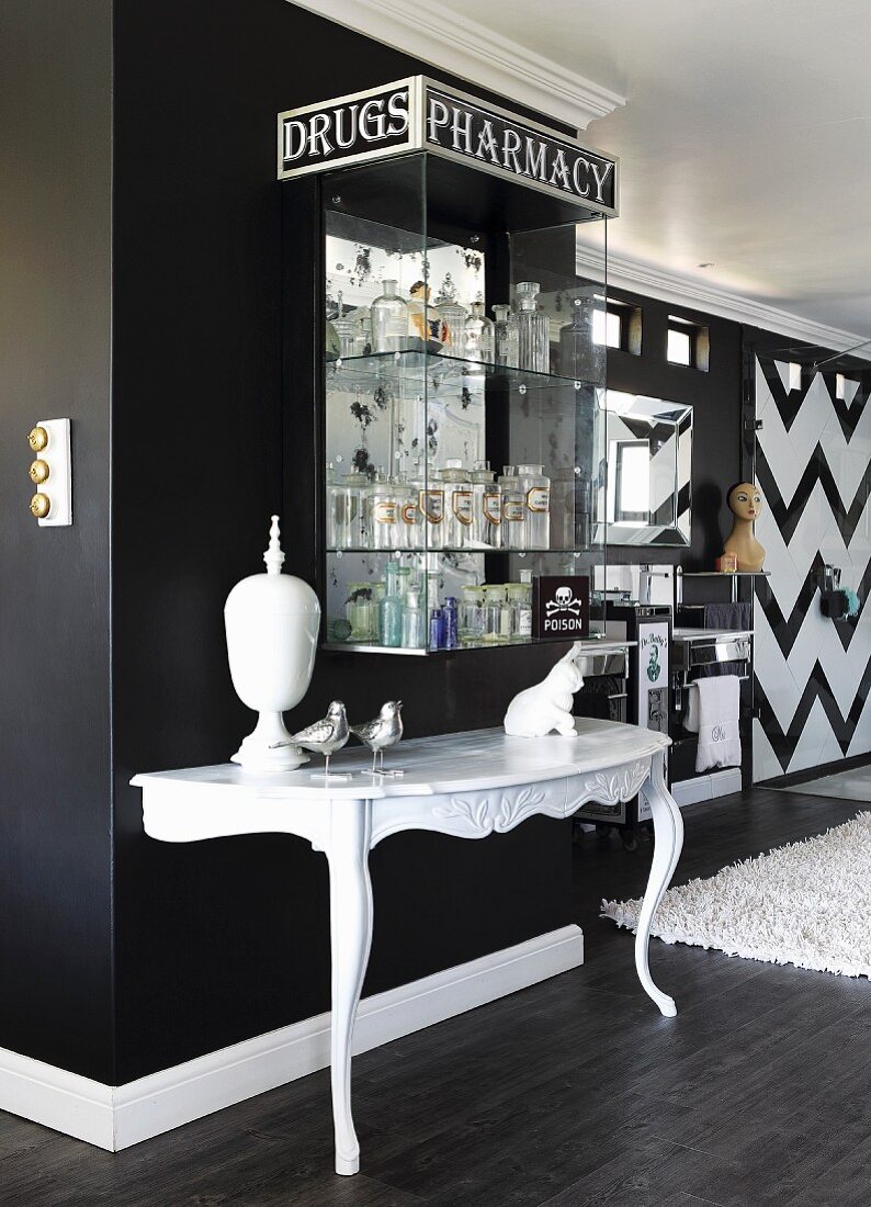 White console table against black wall below collection of apothecaries' bottles in glass case with lettered sign