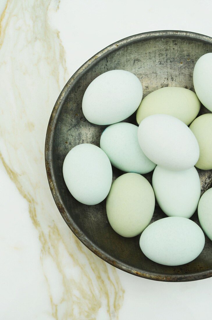A pewter bowl filled with blue eggs on a white marble surface