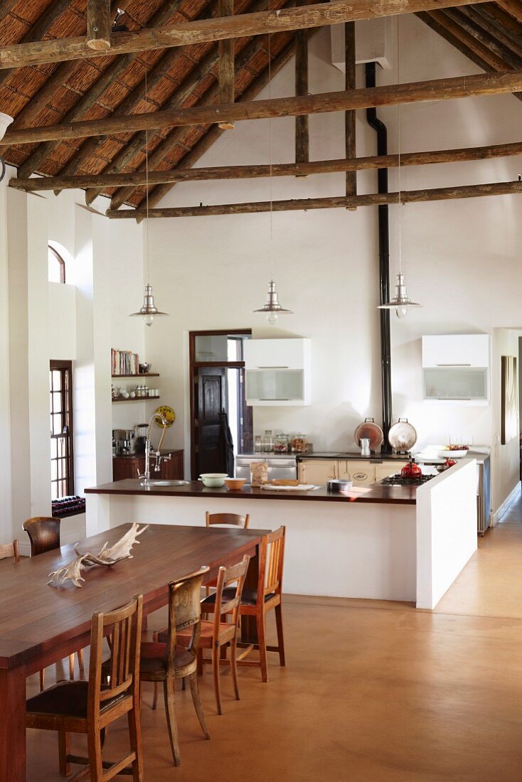 Dining table and various wooden chairs in front of open-plan kitchen with u-shaped counter in high-ceilinged interior with exposed roof structure