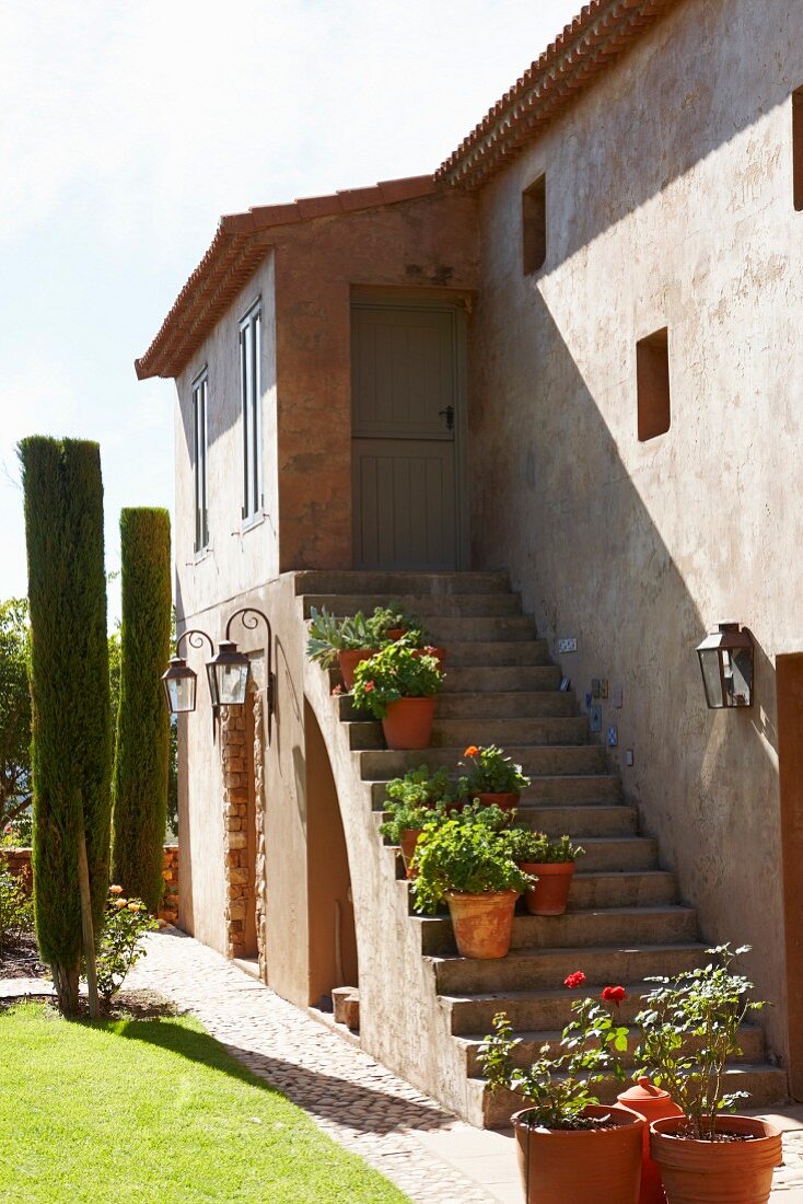 House with potted flowering plants on exterior staircase in sunshine and clipped cypresses in garden