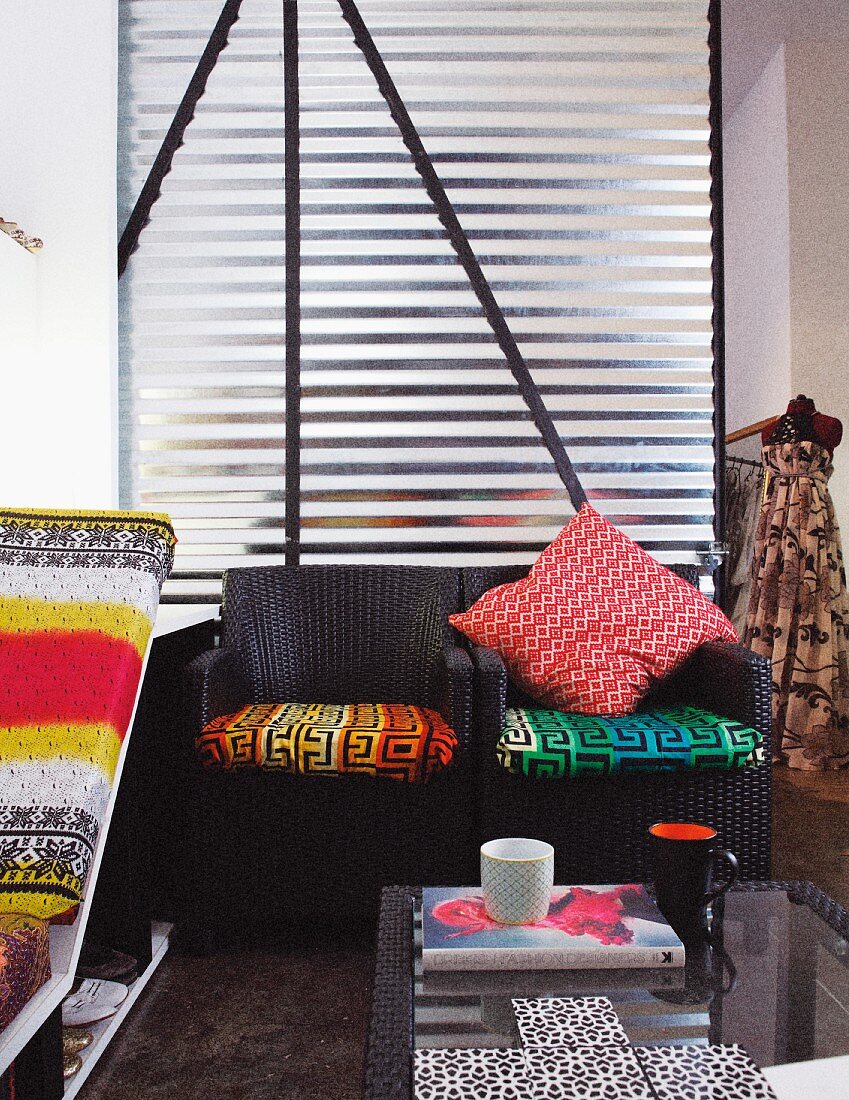 Rattan armchairs with colourful seat cushions against aluminium wall; glass table and chaise with colourful cushions in foreground