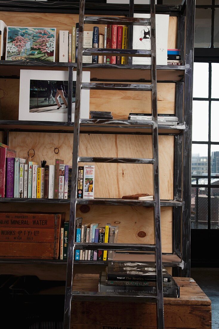Wooden ladder leaning against tall, shabby chic wooden shelves; industrial window with view of cityscape in background