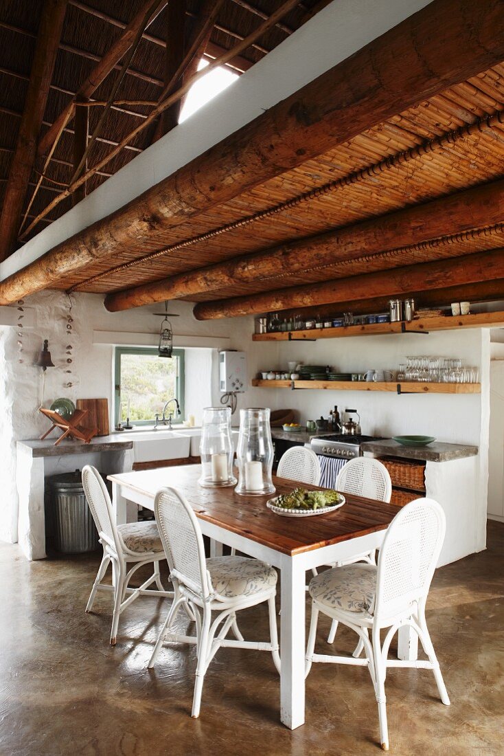 Offener Wohnraum mit einfacher Küche und Holztisch mit weissen Stühlen unter rustikaler Holzbalkendecke