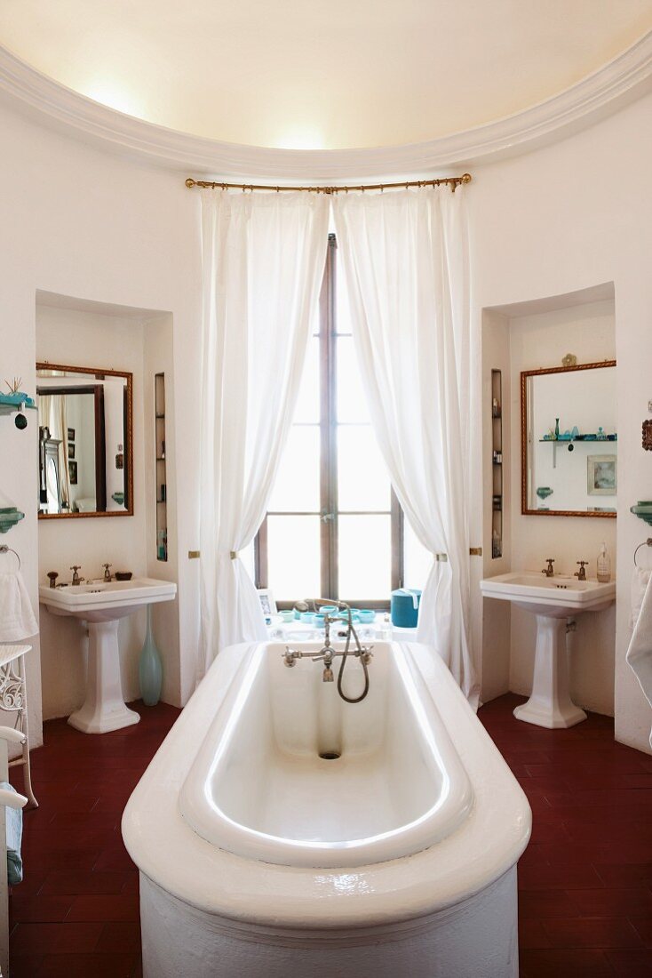 Round bathroom with bathtub in centre of room in front of French windows flanked by twin pedestal washbasins
