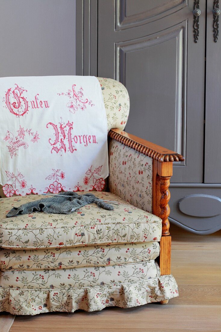 Traditional armchair with floral upholstery and embroidered antimacassar on backrest in front of farmhouse cupboard painted grey