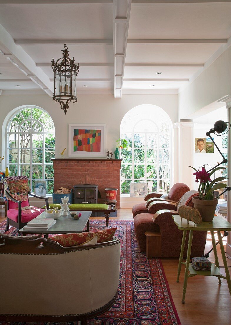 Bright living room with wood burning stove flanked by tall arched windows; seating area in mixture of styles in foreground