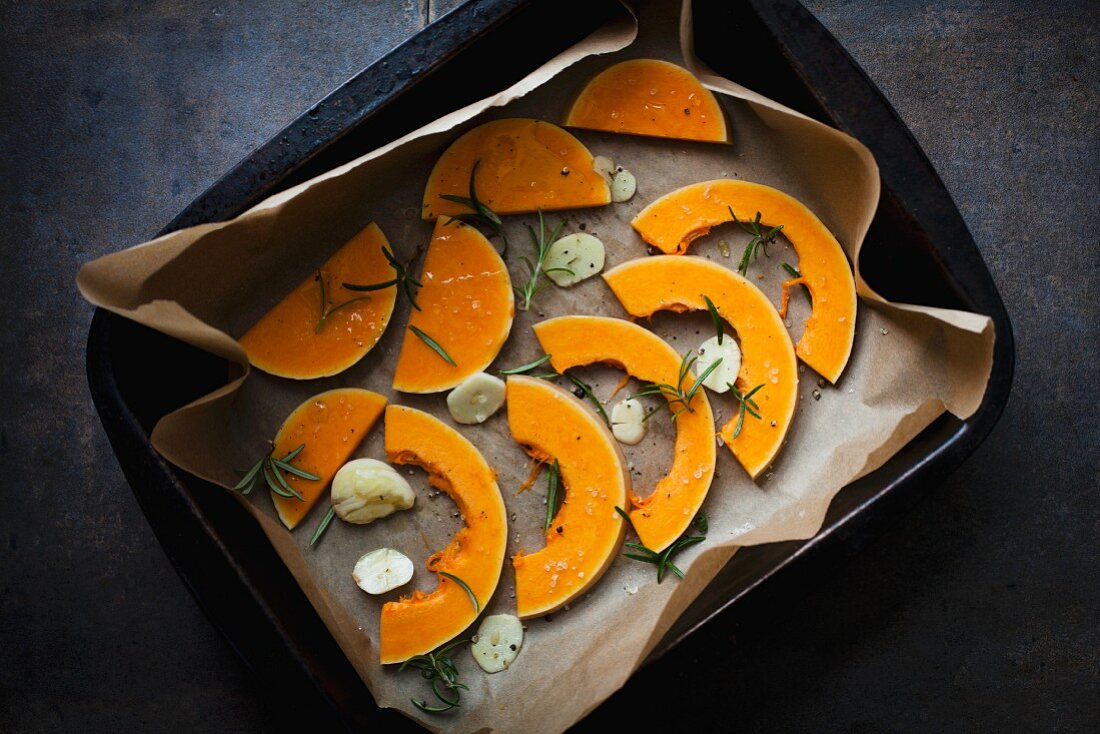 Slices of raw butternut squash with garlic and rosemary in a roasting tin