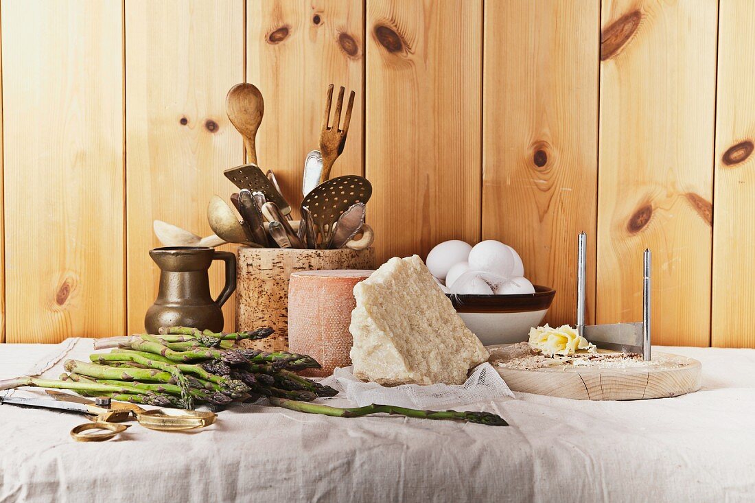 A still life featuring asparagus, cheese, eggs and kitchen utensils on a table