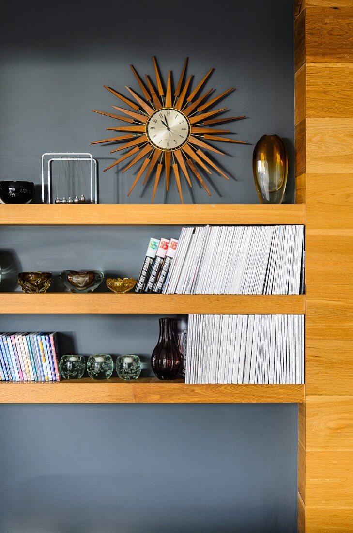 Collection of Murano glass ornaments and books on wooden shelves against grey wall below sunburst-shaped wall clock