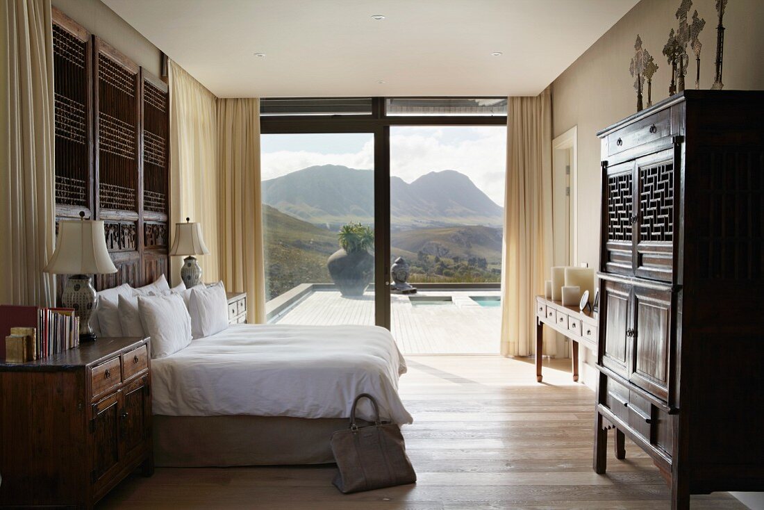 Helles Schlafzimmer mit antiken Holzmöbeln und mit herrlichem Blick auf die Landschaft durch eine raumhohe Glasfront