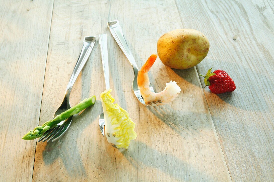 Stillleben mit Spargel, Salatblatt, Garnele, Kartoffel und Erdbeere auf Gabeln