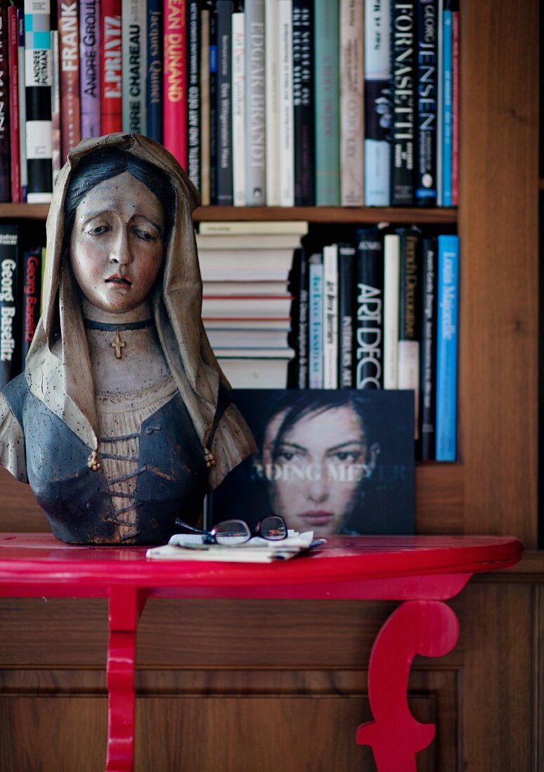 Bemalte, hölzerne Madonnenbüste auf rotem Konsolentisch vor Bücheregal