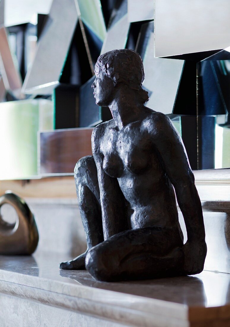 Sitzende Frauenfigur auf Kaminsims; im Hintergrund kubistisches Kunstobjekt mit geneigten Spiegelflächen
