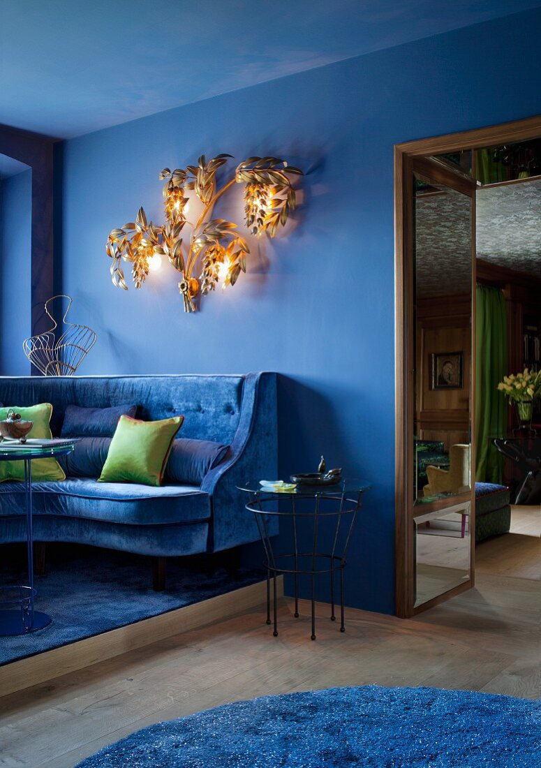 Blauer Salon mit Samtsofa und grünen Dekokissen; französische Clubatmosphäre durch vergoldeten Leuchter aus den 70ern mit floralem Motiv