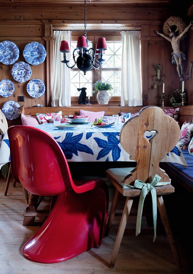 Gedeckter Tisch in Zirbelstube mit Herrgottswinkel und hessischen Fayencen an der Wand; roter Pantonstuhl als Kontrast neben einem Bauernstuhl