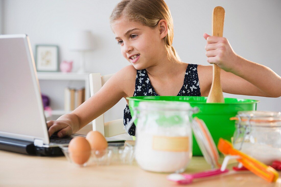 A girl stirring cake mixture and looking at a laptop at the same time