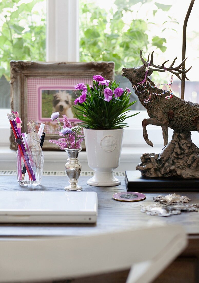 Schreibtisch mit weißem Laptop und pastellfarbenem Blumenschmuck, nostalgische Hirschfigur und gerahmtes Hundefoto im Hintergrund