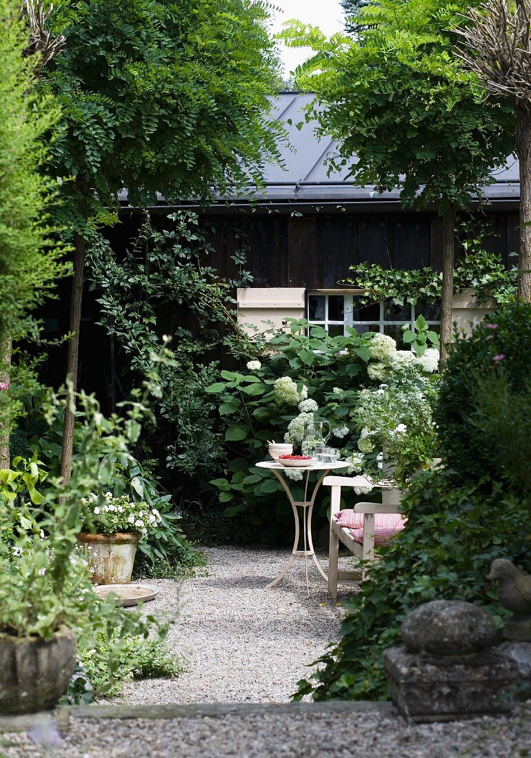 Sonniges Gartenplätzchen - Beistelltisch und Bank zwischen Bäumen auf Kiesboden, im Hintergrund altes Holzhaus