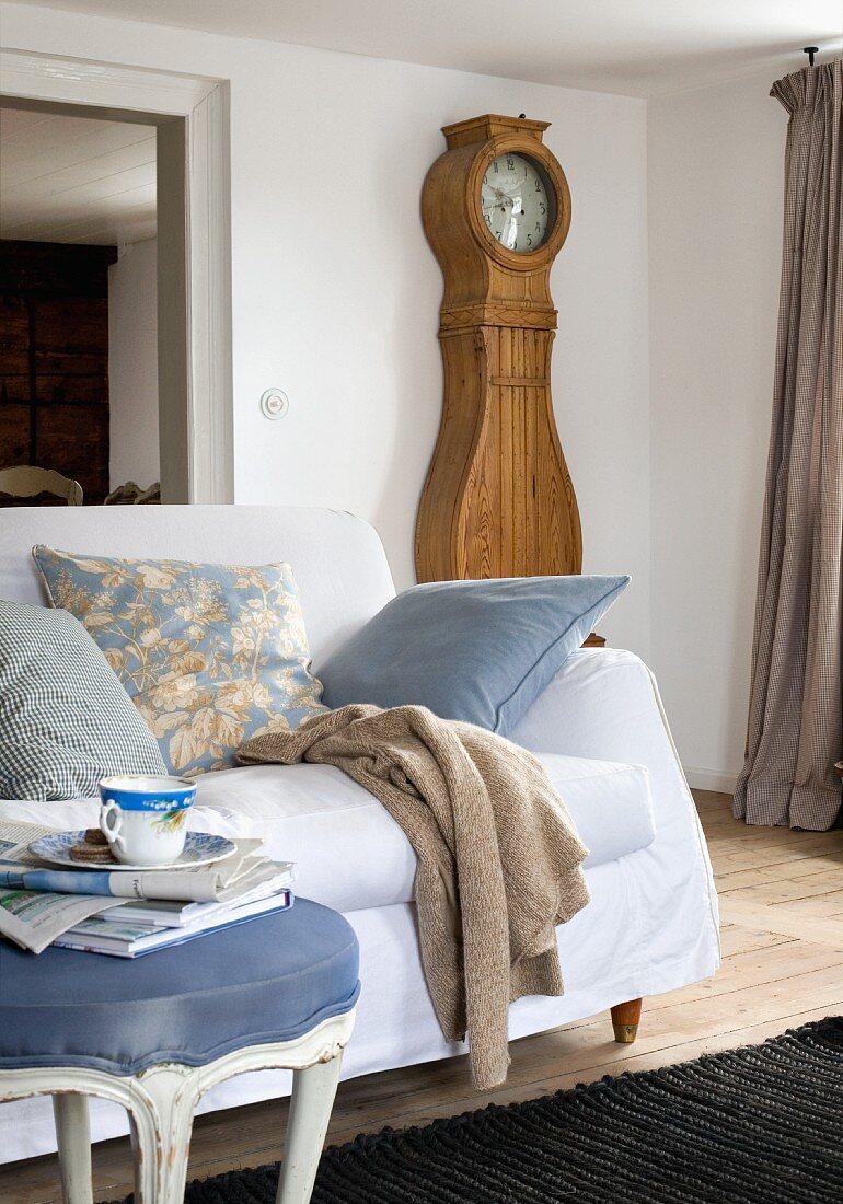 Teacup on footstool in front of couch with white cover and Biedermeier longcase clock in simple interior