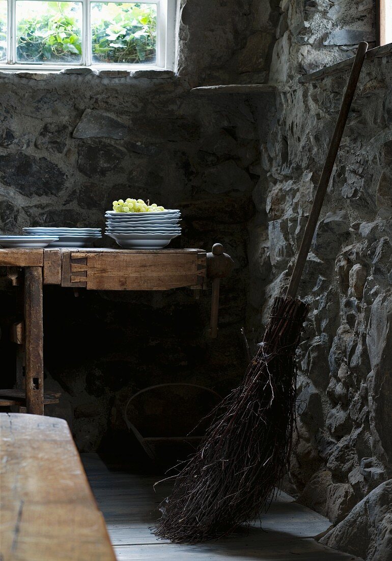 Alter Besen neben Tisch mit Tellerstapel und Trauben im Kellerraum mit Steinmauern