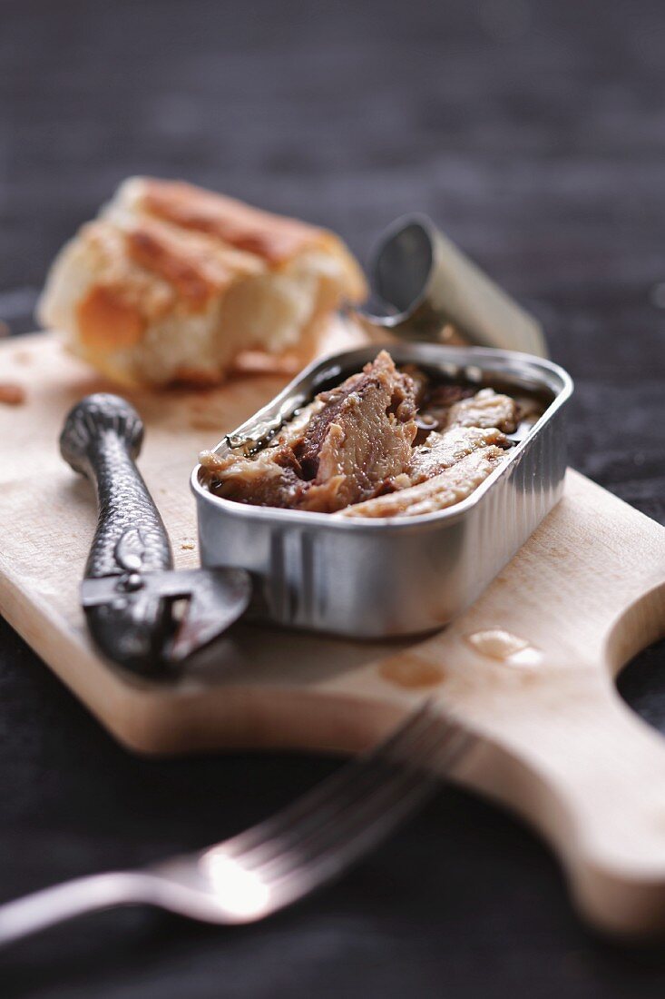 An open can of sardines and a chunk of baguette on a chopping board