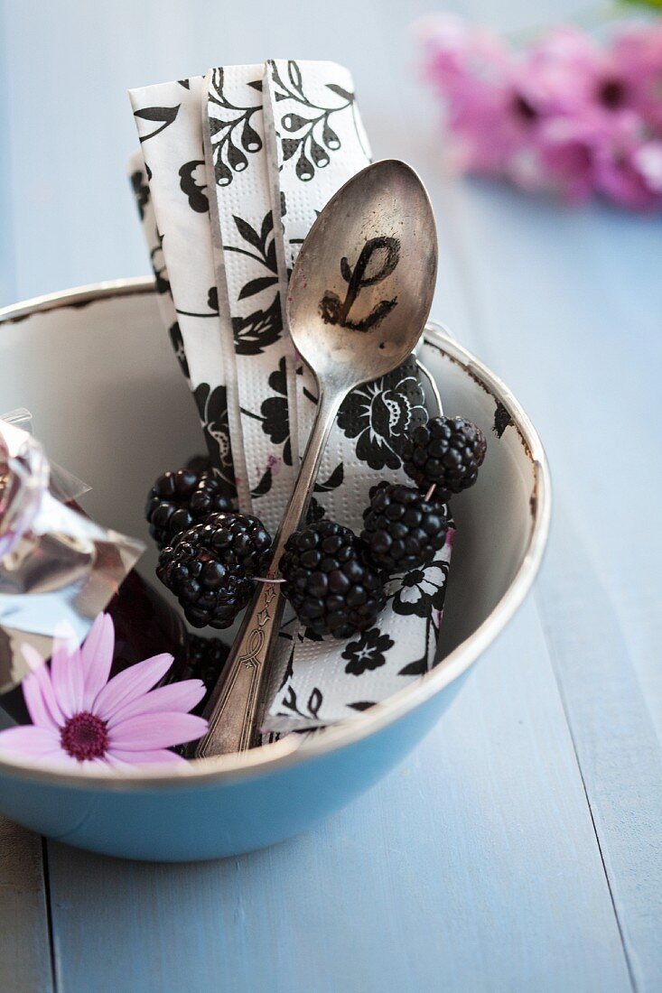 A napkin with a blackberry napkin ring and a spoon with initials in a blue enamel bowl