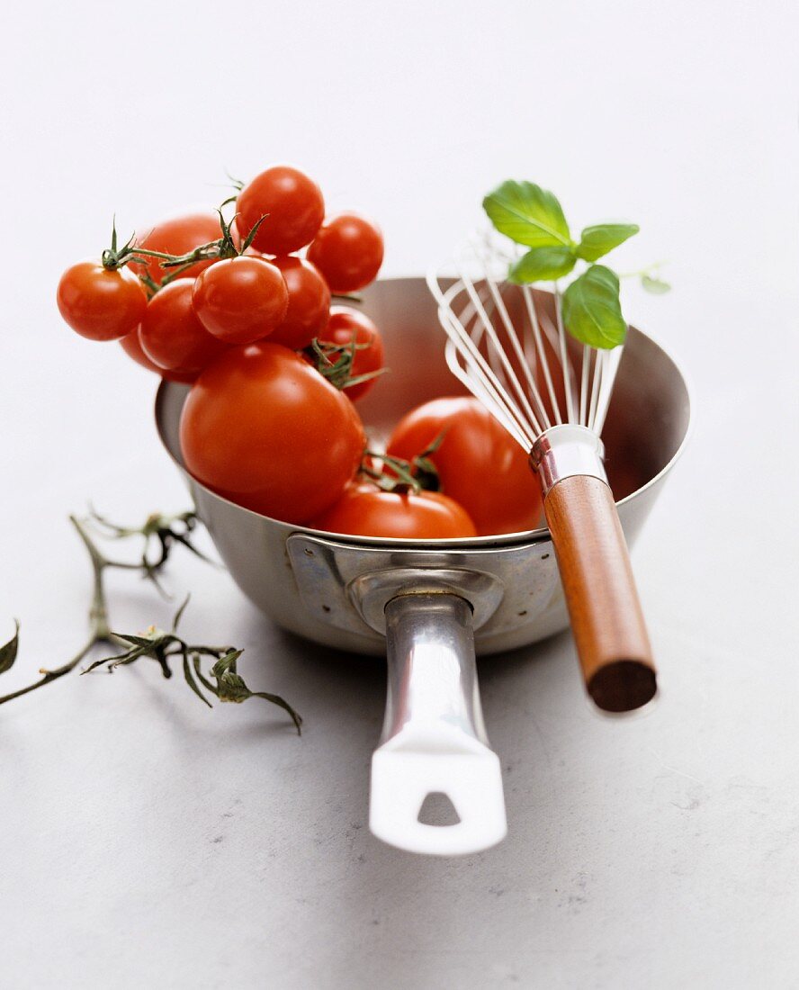 A still life featuring fresh tomatoes, a saucepan and an egg whisk