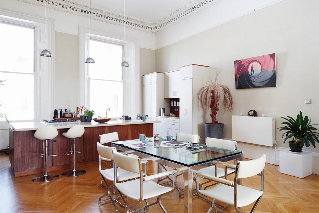 Glass-topped dining table and white leather chairs in front of open-plan kitchen with bar stools at free-standing counter in grand interior